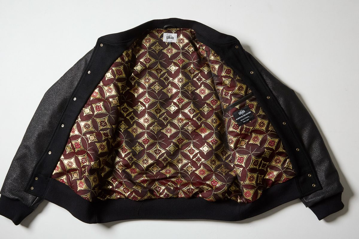 THE URAh のバーシティジャケットを開いて平置きした一着。黒のウールの身頃とレザーの袖の内側に、えんじと金の七宝柄の裏地。